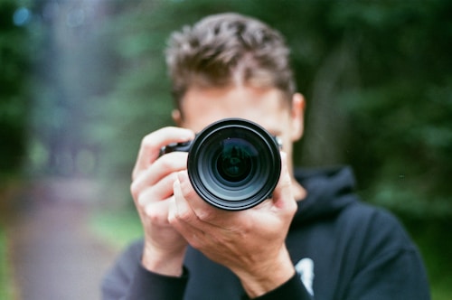 A person is holding a camera with a large lens, pointed directly at the viewer. The background is outdoors, with greenery visible, possibly suggesting a park or forest setting. The focus is on the camera, making the person slightly blurred.