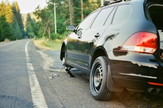 A black car is parked on the side of a highway with a flat tire. The car is jacked up, and the wheel is removed, indicating a tire change in progress. The road extends into the distance lined with lush green trees, and a section of the forest is bathed in golden light.