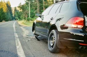 A black car is parked on the side of a highway with a flat tire. The car is jacked up, and the wheel is removed, indicating a tire change in progress. The road extends into the distance lined with lush green trees, and a section of the forest is bathed in golden light.