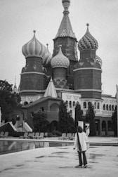 A person stands in front of an ornate building with onion-shaped domes, typical of Russian architecture. The architecture features intricate designs and patterns, lending the structure a historical and cultural significance. Trees are planted around the building, and empty lounge chairs are arranged around a nearby pool.