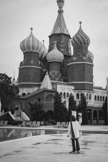 A person stands in front of an ornate building with onion-shaped domes, typical of Russian architecture. The architecture features intricate designs and patterns, lending the structure a historical and cultural significance. Trees are planted around the building, and empty lounge chairs are arranged around a nearby pool.
