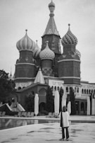 A person stands in front of a Russian-style building with distinctive onion domes, capturing a gray and white monochrome scene. The architecture features intricate patterns and spires, surrounded by trees and umbrellas.