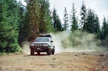 An off-road vehicle driving on a dirt path surrounded by dense, tall evergreen trees. Dust clouds are visible behind the vehicle, suggesting movement and speed. The outdoor setting appears rugged and natural.