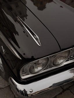 A close-up angle of a vintage car's front section, highlighting the glossy black paint and chrome detailing. The image captures the car's iconic headlight design with a reflective, polished surface. A faint reflection of the surrounding street and people is visible on the surface of the car.