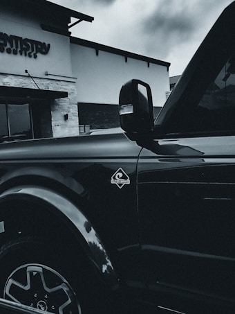 A black vehicle is parked in front of a building with signage that reads 'Dentistry.' The vehicle's side view mirror and part of the door are visible, along with the 'Outer Banks' emblem. The architecture of the building includes stone and brick elements.