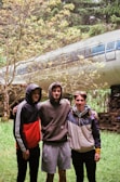 Three young men stand closely together with arms around each other's shoulders in a lush forest setting. Behind them, an old airplane fuselage is nestled among the trees, giving the scene an adventurous and slightly mysterious vibe.