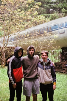 Three young men stand closely together with arms around each other's shoulders in a lush forest setting. Behind them, an old airplane fuselage is nestled among the trees, giving the scene an adventurous and slightly mysterious vibe.