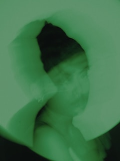 An abstract image with a predominantly green tint featuring a person with a blurred or double-exposed effect. The person appears to have dark hair and is positioned centrally within the frame.