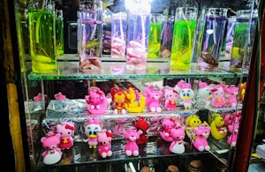 The image displays a glass shelf with two levels. The top level contains several tall glass bottles filled with colorful liquids, possibly for decorative purposes, featuring items such as preserved plants and shells. The bottom level showcases a variety of small, colorful figurines and keychains, many of which are designed in a cartoon style with bright colors like pink and yellow.