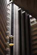 An interior view of a modern building featuring a sleek, vertical elevator shaft with glass elevators and balconies. The architecture showcases geometric patterns with a mix of concrete and metal elements. Soft lighting adds a warm ambiance to the scene.
