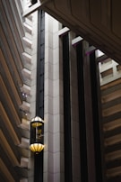 An interior view of a modern building featuring a sleek, vertical elevator shaft with glass elevators and balconies. The architecture showcases geometric patterns with a mix of concrete and metal elements. Soft lighting adds a warm ambiance to the scene.