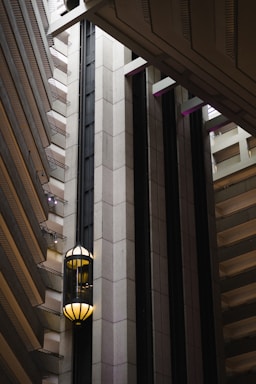 An interior view of a modern building featuring a sleek, vertical elevator shaft with glass elevators and balconies. The architecture showcases geometric patterns with a mix of concrete and metal elements. Soft lighting adds a warm ambiance to the scene.