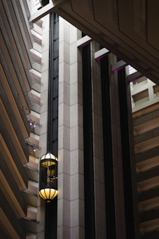 An interior view of a modern building featuring a sleek, vertical elevator shaft with glass elevators and balconies. The architecture showcases geometric patterns with a mix of concrete and metal elements. Soft lighting adds a warm ambiance to the scene.