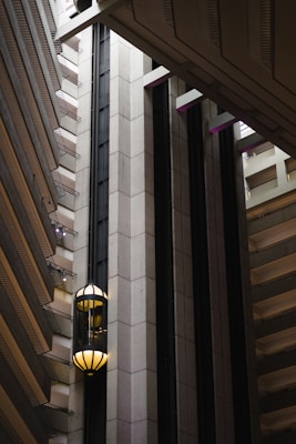 An interior view of a modern building featuring a sleek, vertical elevator shaft with glass elevators and balconies. The architecture showcases geometric patterns with a mix of concrete and metal elements. Soft lighting adds a warm ambiance to the scene.