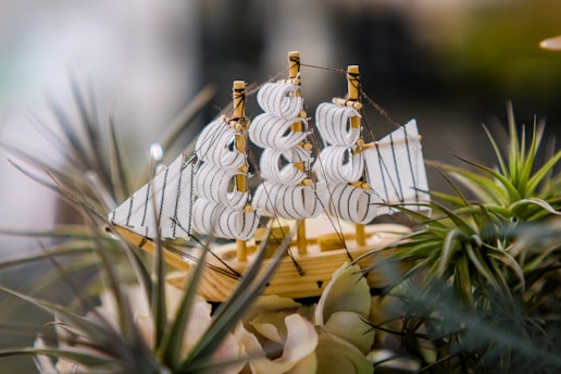 A detailed model of a sailing ship with intricate white sails and ropes is surrounded by green and beige decorative plants. The wooden hull of the ship contrasts with the lush greenery around it.