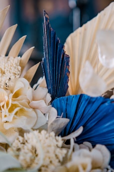 A close-up of an arrangement of dried and artificial flowers, featuring a mix of soft, cream-colored petals and bold, blue palm-shaped leaves. The composition includes various textures and shapes, creating an intricate and artistic display.