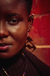 A close-up of the right side of a person’s face with rich, dark skin, adorned with a large geometric earring and multiple piercings. The background is a textured, warm red wall. The person wears a black top and a multicolored beaded necklace.