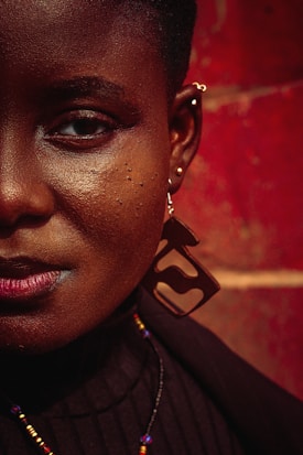 A close-up of the right side of a person’s face with rich, dark skin, adorned with a large geometric earring and multiple piercings. The background is a textured, warm red wall. The person wears a black top and a multicolored beaded necklace.