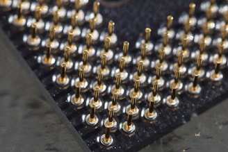 A close-up view of a grid of metallic pins protruding from a black surface, likely part of a computer processor. The pins are uniformly arranged and appear to be gold-plated, reflecting light in a bright contrast against the dark background.