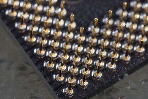 A close-up view of a grid of metallic pins protruding from a black surface, likely part of a computer processor. The pins are uniformly arranged and appear to be gold-plated, reflecting light in a bright contrast against the dark background.