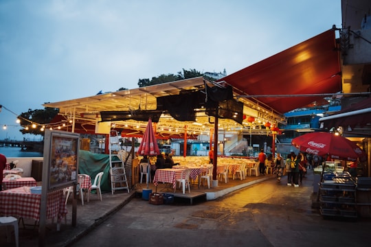 A bustling outdoor market or dining area with tables covered in red and white checkered cloths under a roof with bright lights. Several people are gathered around the tables, and umbrellas displaying a popular beverage brand provide cover. The setting is in the evening with a clear sky.