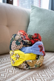 A collection of colorful, intricately designed headbands is stacked on a patterned surface. The headbands feature various designs, including floral patterns, sequins, and beads, with prominent colors of red, blue, and yellow.