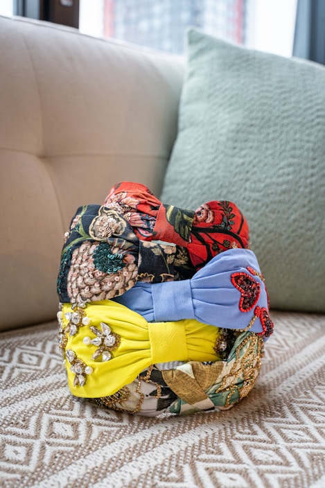A collection of colorful, intricately designed headbands is stacked on a patterned surface. The headbands feature various designs, including floral patterns, sequins, and beads, with prominent colors of red, blue, and yellow.