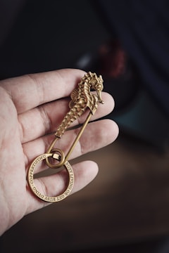 A hand is holding a brass-colored safety pin featuring a seahorse design. Attached to the pin is a circular metal ring inscribed with words.
