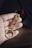 A hand is holding a brass-colored safety pin featuring a seahorse design. Attached to the pin is a circular metal ring inscribed with words.
