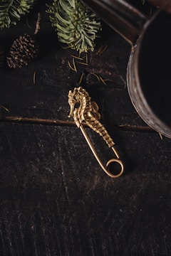 A golden seahorse-shaped decorative pin is placed on a dark, rustic wooden surface. Surrounding elements include green pine needles and a pine cone, suggesting a forest or woodland theme.