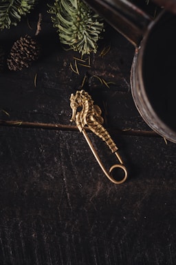 A golden seahorse-shaped decorative pin is placed on a dark, rustic wooden surface. Surrounding elements include green pine needles and a pine cone, suggesting a forest or woodland theme.