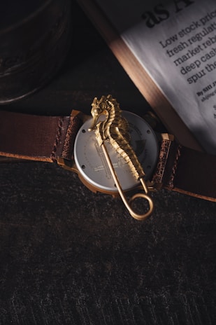 A watch with a leather strap lies on a dark surface, featuring a prominent golden seahorse decoration across its back. To the side, a partially visible newspaper adds an element of sophistication.