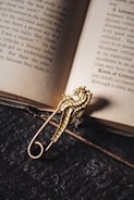 An open book lays on a dark, textured surface. A metallic, gold-colored seahorse-shaped brooch is placed on the book, casting a shadow.