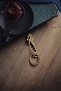 A gold seahorse brooch with a circular loop lies on a wooden surface. Nearby, there is a dark, round candle holder with a red candle, slightly melted. The edge of a textured, dark blue book is partially visible.
