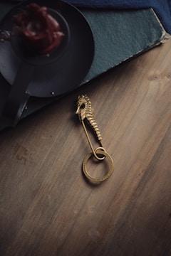 A gold seahorse brooch with a circular loop lies on a wooden surface. Nearby, there is a dark, round candle holder with a red candle, slightly melted. The edge of a textured, dark blue book is partially visible.