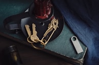 A collection of objects arranged on a dark-colored surface, including two antique-style keys attached to a seahorse-shaped pendant, a metallic USB stick, a candle holder with a red candle, and the corner of a hardcover book. A textured blue fabric drapes nearby, adding a rich color contrast.