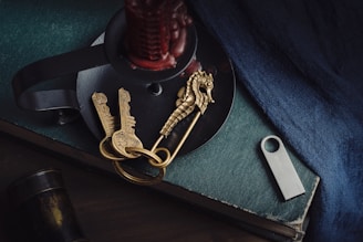 A collection of objects arranged on a dark-colored surface, including two antique-style keys attached to a seahorse-shaped pendant, a metallic USB stick, a candle holder with a red candle, and the corner of a hardcover book. A textured blue fabric drapes nearby, adding a rich color contrast.