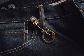 A close-up view of denim jeans featuring detailed yellow stitching and a glossy, metallic horse-shaped pin. The pin is attached near the pocket, adding a unique decorative element.