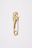 A gold safety pin featuring an ornate seahorse design, positioned vertically against a plain, light gray background. The pin is intricately detailed, capturing the textured body and curling tail of the seahorse, which forms the upper part of the pin.