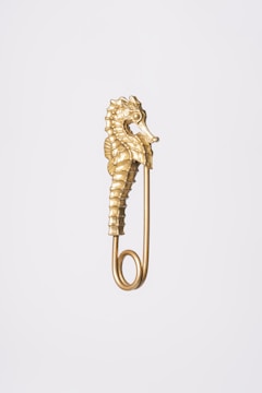 A gold safety pin featuring an ornate seahorse design, positioned vertically against a plain, light gray background. The pin is intricately detailed, capturing the textured body and curling tail of the seahorse, which forms the upper part of the pin.