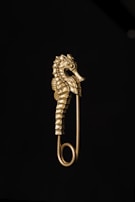 A gold-colored safety pin features an intricately designed seahorse as its decorative element. The seahorse is detailed, showcasing its distinctive texture and contours against a dark background.
