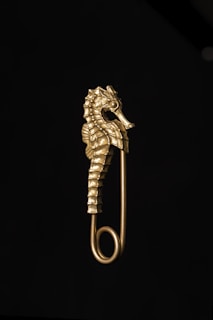 A gold-colored safety pin features an intricately designed seahorse as its decorative element. The seahorse is detailed, showcasing its distinctive texture and contours against a dark background.