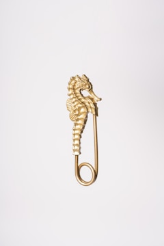 A gold-colored seahorse figurine integrated into the design of a safety pin, positioned against a plain white backdrop.