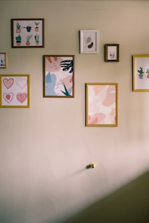 A wall adorned with several framed artworks, featuring abstract designs and illustrations of plants. The frames vary in size and color, and are neatly arranged, creating an aesthetically pleasing display.