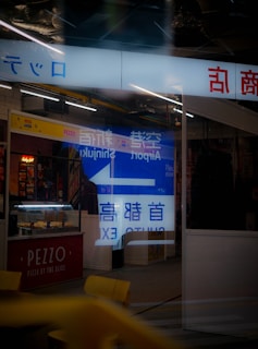 A storefront showcasing a brightly lit interior with reflective glass displaying text and signage in multiple languages. The glass reflects neon lights and displays advertisements, including a sign for 'PEZZO Pizza by the Slice.' The overall composition features urban elements and marketing signage.