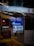 A storefront showcasing a brightly lit interior with reflective glass displaying text and signage in multiple languages. The glass reflects neon lights and displays advertisements, including a sign for 'PEZZO Pizza by the Slice.' The overall composition features urban elements and marketing signage.