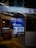 A storefront showcasing a brightly lit interior with reflective glass displaying text and signage in multiple languages. The glass reflects neon lights and displays advertisements, including a sign for 'PEZZO Pizza by the Slice.' The overall composition features urban elements and marketing signage.