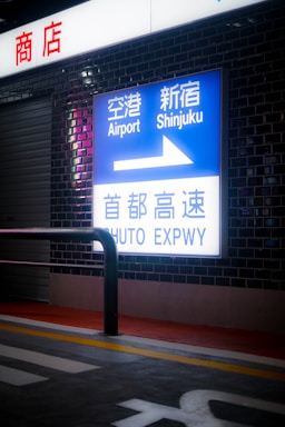 A brightly lit sign mounted on a dark brick wall displaying directions to the Airport and Shinjuku, along with an arrow. The sign includes text in both Japanese and English. Above the sign, another light box with red Japanese characters is present. A metal railing and part of a roadway are visible at the bottom, with yellow and white lines on the pavement.