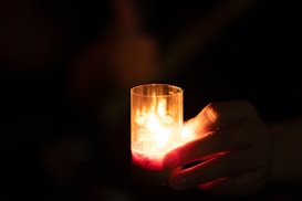 A hand gently holds a lit candle in a dimly lit environment, casting a warm and soft glow. The flame flickers inside a clear glass container, creating an intimate and serene ambiance.