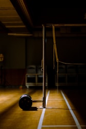 An indoor sports hall with a badminton net stand prominently positioned in the center. The scene is dimly lit, creating a dramatic contrast between the bright wooden floor and the dark background. A lone shuttlecock carrier with wheels is visible near the base of the net post.